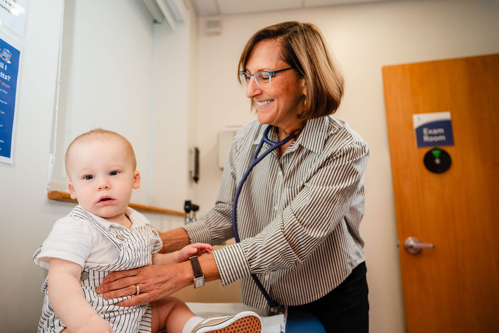 a smiling woman holding a stethoscope to the chest of a young infant child